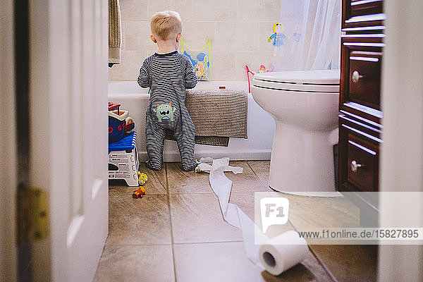Ein kleiner Junge steht mit einer ausgerollten Toilettenpapierrolle in einem Badezimmer.