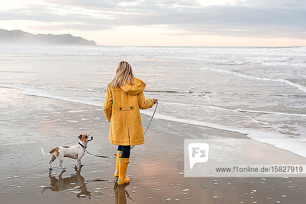 Tween in gelbem Mantel geht mit Hund am Strand in Neuseeland spazieren