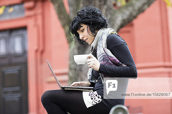 young woman reading in a laptop with a coffee cup outside