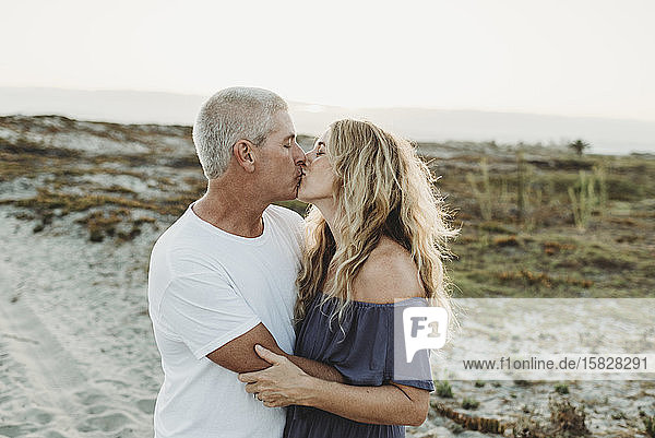 Husband and wife embracing and kissing at beach during sunset