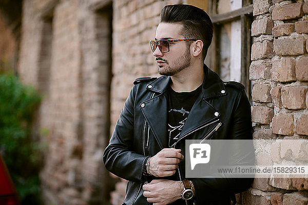 Trendy Man with sunglasses in a jacket standing by a brick wall.
