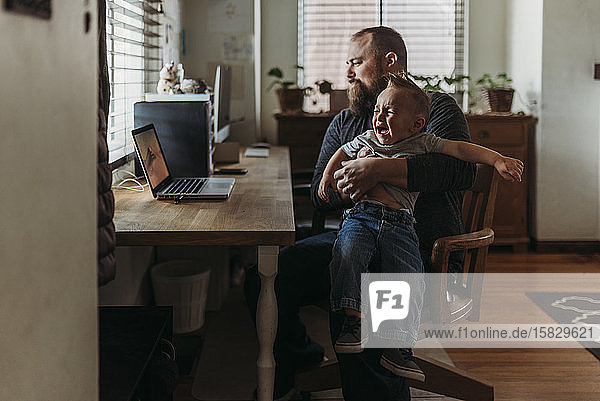Dad working from home with one year old boy crying in his lap