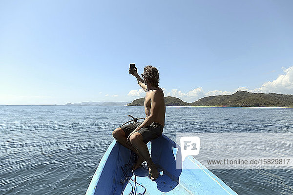 Rückenansicht eines Mannes mit Mobiltelefon  der am Bug eines Bootes sitzt