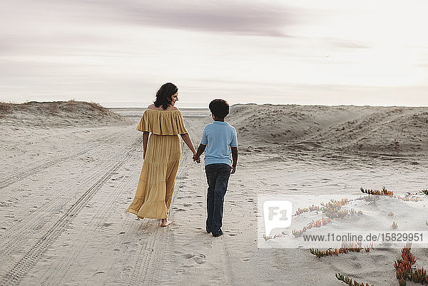Young mother and school-aged son walking at beach against cloudy sky
