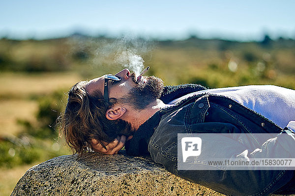 Porträt eines jungen tätowierten Mannes  der auf einem Felsen liegend raucht