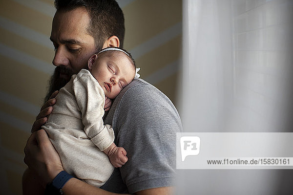 Father embracing sleeping newborn daughter on shoulder at home