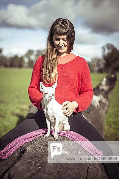 Frau in knallrotem Pullover sitzt mit kleinem weißen Hund auf einem Baumstamm auf dem Bauernhof