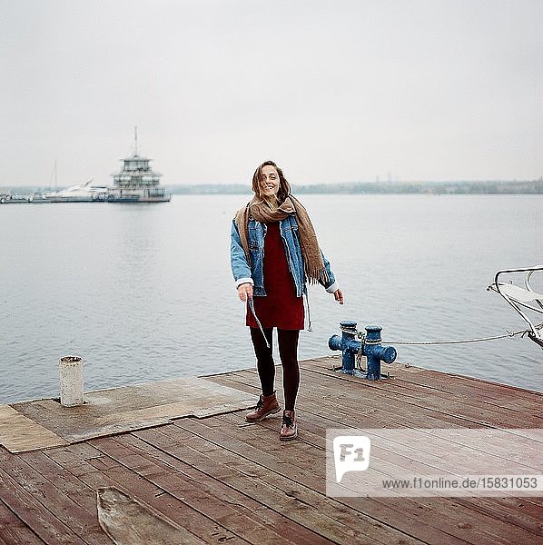 Stylish woman in red dress and blue jacket standing on old wooden pier