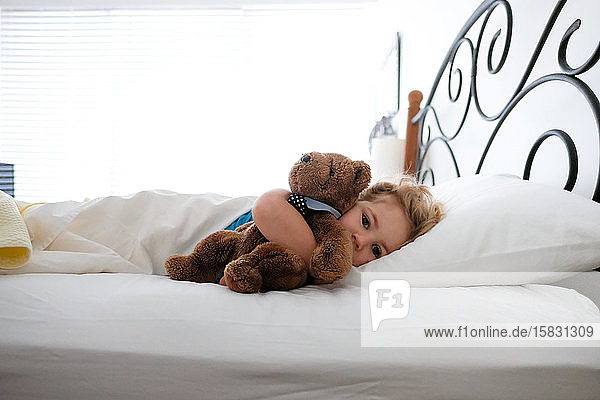 kleines Mädchen liegt im Bett und kuschelt mit einem Teddybär und schaut in die Kamera
