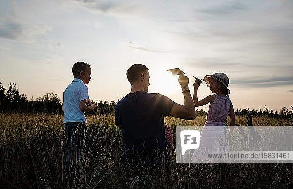 Vater spielt mit Papierflugzeugen mit Sohn und Tochter bei Sonnenuntergang