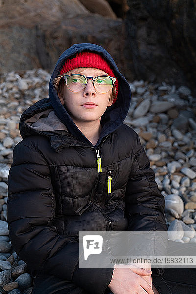 Junge  der mit einer Brille am Strand aus Steinen sitzt und den Sonnenuntergang betrachtet