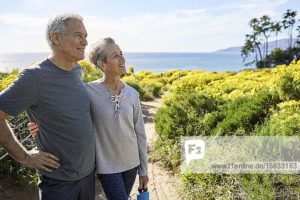 Lächelndes älteres Paar schaut weg  während es an einem sonnigen Tag auf dem Weg gegen den Himmel steht