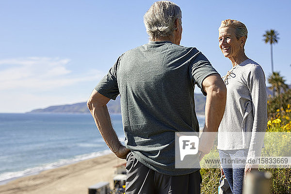 Happy senior couple talking on cliff at beach against sky during sunny day