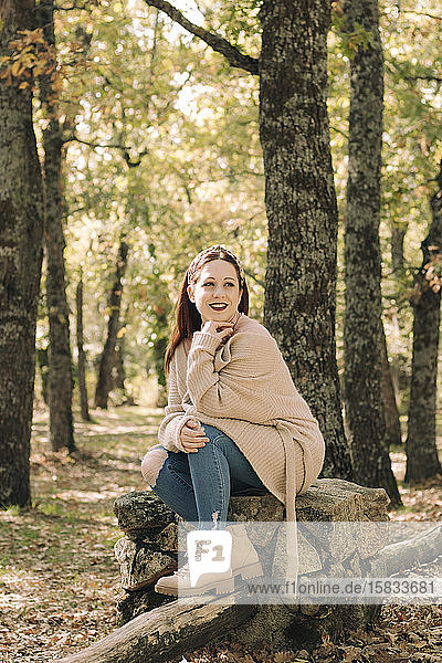 Lächelnde junge Frau mit braunen Haaren sitzt im Herbst in einem Wald
