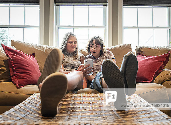 Zwei Mädchen im Teenageralter sitzen auf einer Couch mit den Füßen nach oben und schauen auf ihr Handy.