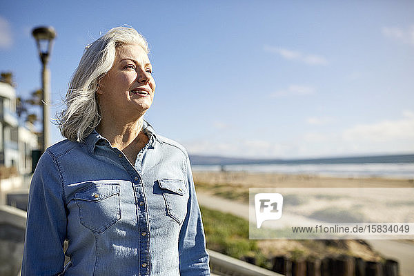 Nachdenklich lächelnde ältere Frau steht am Strand von Manhattan am sonnigen Tag am Himmel