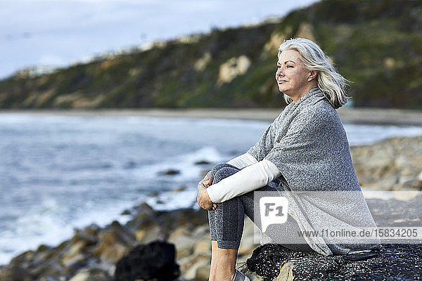 Side view of thoughtful senior woman sitting on rock at Manhattan Beach during sunset