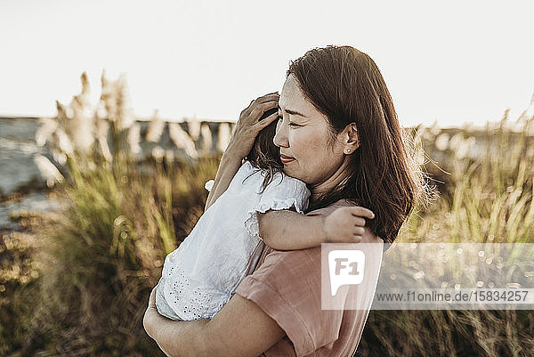 Mitteansicht einer liebenden Mutter  die ihre kleine Tochter bei Sonnenuntergang am Strand umarmt