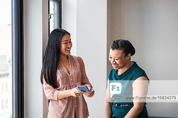 Smiling businesswoman showing mobile phone to female colleague by window at office