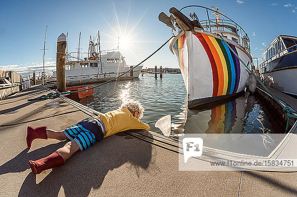 Little kid with net lying on dock next to boat with rainbow