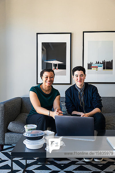Porträt von selbstbewusst lächelnden Geschäftskollegen mit Laptop am Tisch sitzend im Büro