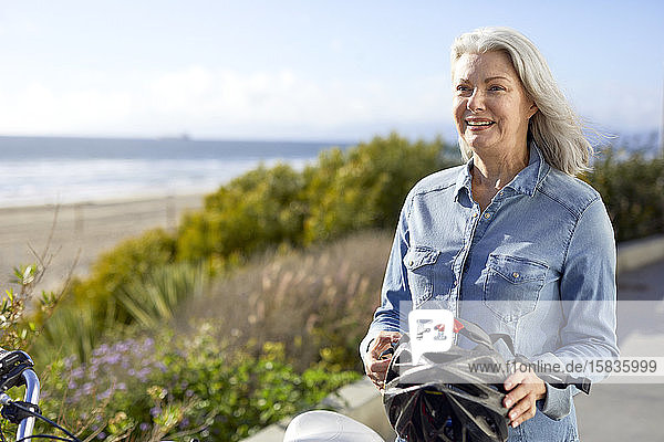 Lächelnde ältere Frau mit Fahrradhelm  während sie an einem sonnigen Tag auf der Straße gegen den Himmel steht