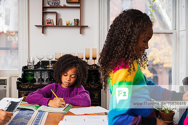 Mädchen macht Hausaufgaben  während die Schwester im Wohnzimmer am Tisch steht