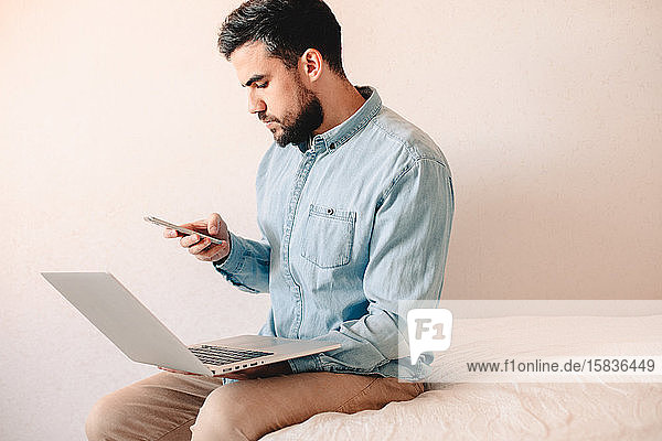Mann hält Laptop und Smartphone in der Hand  während er zu Hause auf dem Bett sitzt