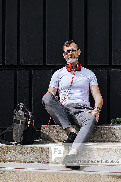 Bearded mature man sitting on steps with headphones while relaxing on a sunny day