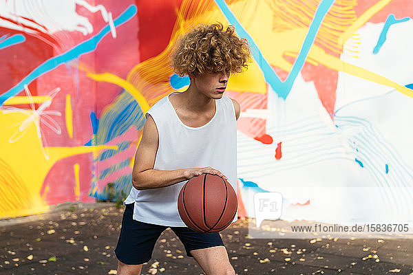 Men with curvy hair and white t-shirt play basketball outside in a court with graffiti