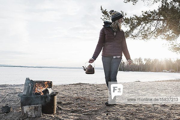 Frau  die in Schweden mit einem Teekessel zum Lagerfeuer an einem Strand geht