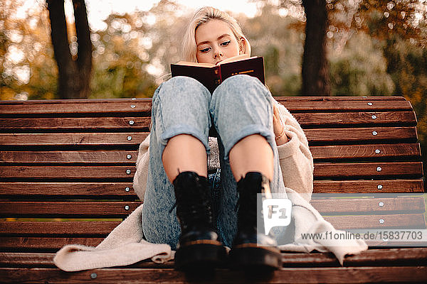 Teenager-Mädchen liest im Herbst auf einer Parkbank sitzend ein Buch