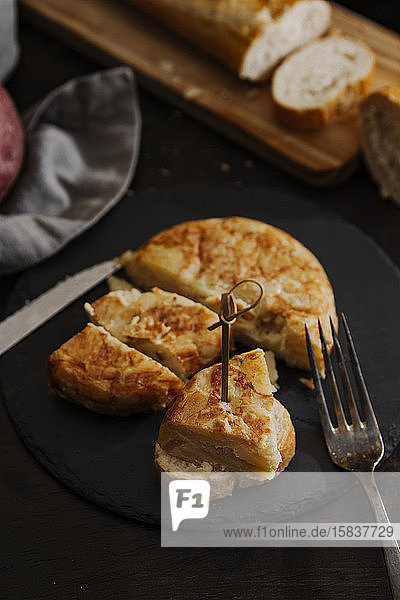 Eine Scheibe spanisches Kartoffelomelett  serviert auf einer Brotscheibe und einer alten silbernen Gabel. Die Aufnahme wurde aus einem hohen Winkel mit Brot in einem hölzernen Schneidebrett im Hintergrund aufgenommen.