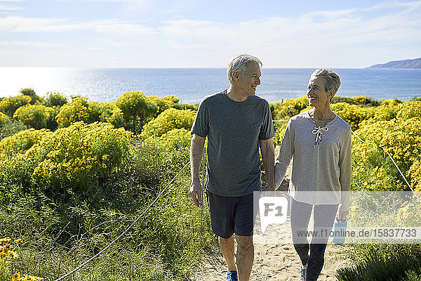 Glückliches älteres Ehepaar hält sich an der Hand  während es am sonnigen Tag auf einem Pfad inmitten von Pflanzen auf einer Klippe am Meer gegen den Himmel läuft
