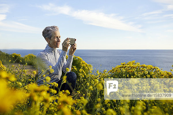 Ältere Frau fotografiert mit einem Smartphone  während sie am Meer vor Pflanzen gegen den Himmel kauert