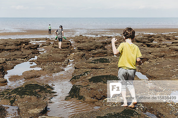 Boy walking over rocky seashore towards sea with family