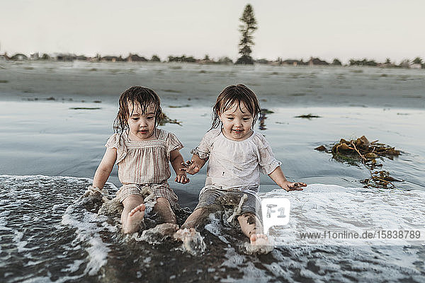 Front view of toddler sisters sitting and splashing in water at beach