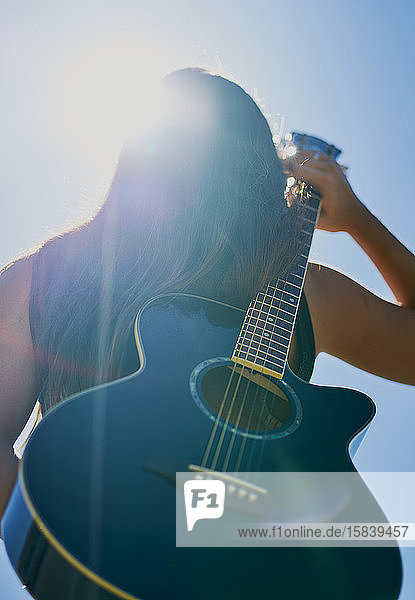 Eine weibliche Teenager-Gitarristin hält sich eine Akustikgitarre auf dem Rücken vor einen blauen Himmel