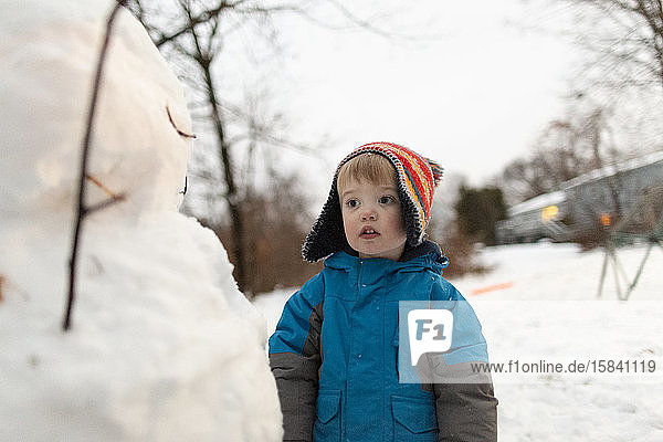 Mid shot of toddler boy wearing knit hat standing looking at snowman
