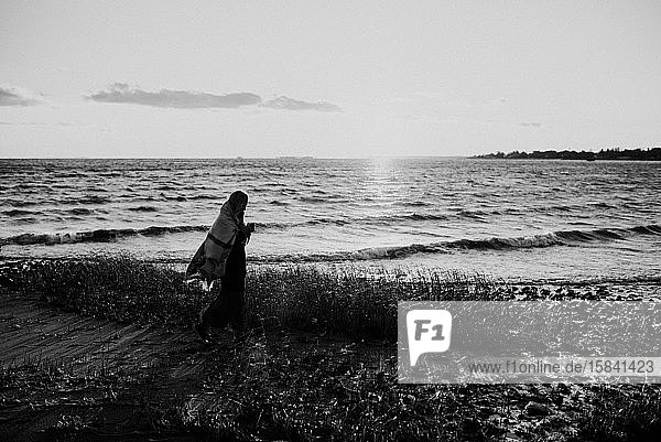 Frau  die bei Sonnenuntergang in einer Decke am Strand spazieren geht