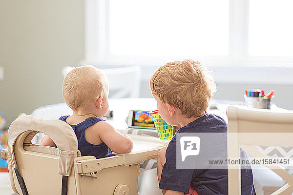Rückansicht eines Mädchens und eines kleinen Bruders beim Telefonieren am Tisch im Haus