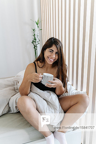 Junges Mädchen sitzt auf dem Bett und hält lächelnd eine Tasse Kaffee