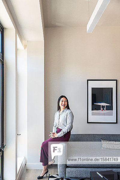 Glückliche Unternehmerin schaut weg  während sie mit gefalteten Händen auf dem Sofa an der Wand im Büro sitzt