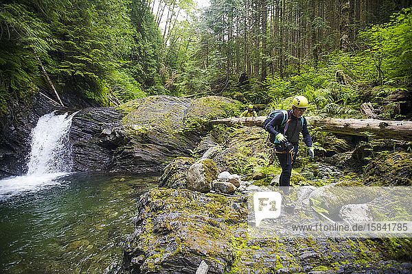Wanderer wandert über felsiges Gelände neben einem Wasserfall in dichtem Wald.