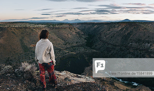 Young woman standing on cliff looking out over canyon at sunset