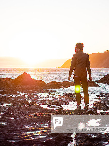 Man standing on rocky shore facing ocean with sun setting