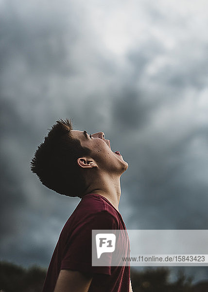 Teenager-Junge  der mit weit geöffnetem Mund in den dunklen Wolkenhimmel schaut.