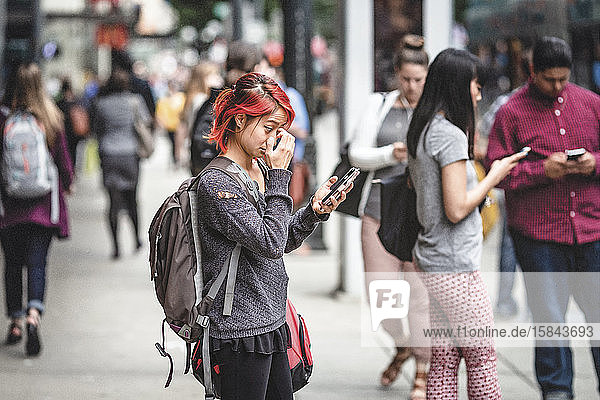 Frau mit rot gefärbten Haaren schaut Handy auf überfüllter Straße an