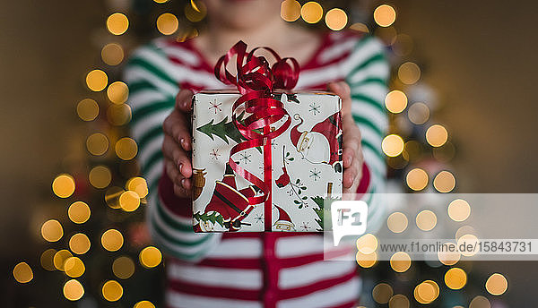 Child in pyjamas holding Christmas gift in front of lights on tree.