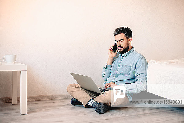 Man talking on smart phone while using laptop computer sitting at home